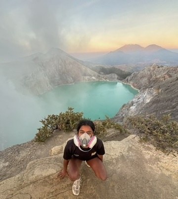 Brittany in a gas mask, crouched above a turquoise crater lake at sunrise